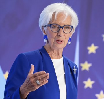 An older woman with short white hair and blue glasses, wearing a blue blazer and statement earrings, speaking at a podium with the European Union flag in the background.