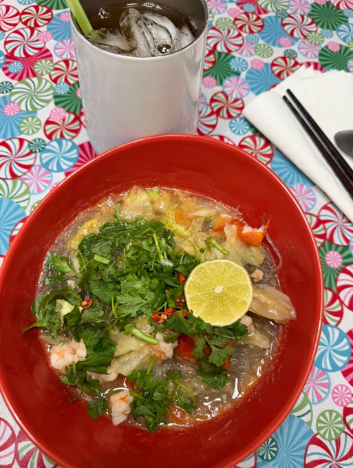 A red bowl of noodle soup topped with herbs, shrimp, and a lemon slice sits beside a metal cup of iced drink on a colorful floral tablecloth.