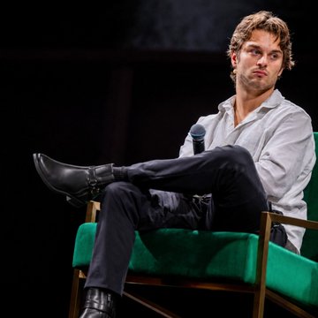 The image features a man sitting in a green chair, with his legs crossed and looking off to the side. The background appears to be a dark room or stage setting. There are two instances of the word "here" in the image, which could potentially be part of a caption or description related to the post about Dior ankle boots.