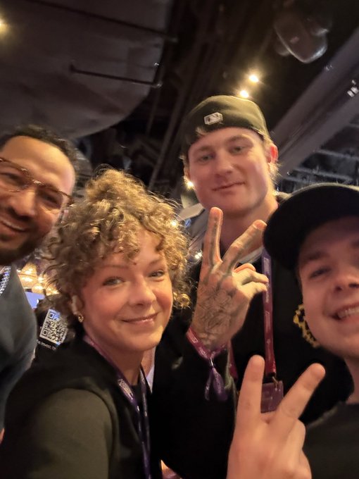 A group selfie of four people at what appears to be a conference or event, with one person making a peace sign and wearing a cap.