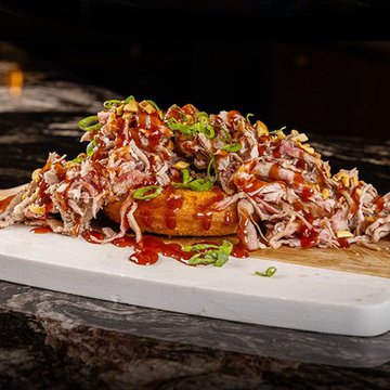 A close-up photograph of a sandwich or toast topped with pulled meat, drizzled with red sauce and garnished with green onions, served on a white cutting board.