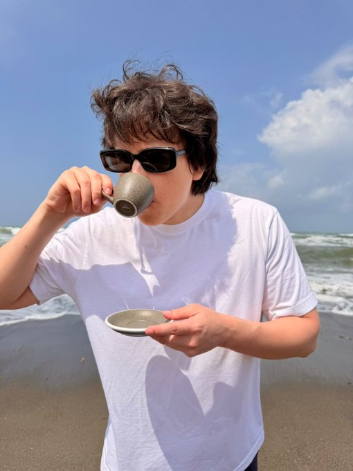 A young man with messy dark hair and sunglasses drinks from a speckled mug while holding a saucer, standing on a sandy beach under a blue sky.