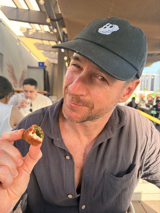 A man in a gray button-up shirt and a black baseball cap smiles while holding a small fried food item topped with a green sauce, seated at what appears to be an outdoor food market.