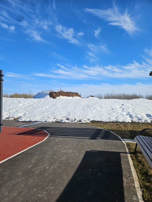 An outdoor winter scene showing a path with red and black pavement, surrounded by snow-covered ground under a bright blue sky with wispy clouds.