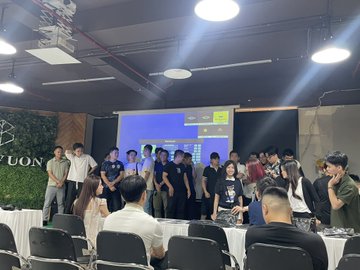 A group of people gathered in a modern indoor venue, some posing for a photo in front of a large digital screen while others sit at tables in the foreground.