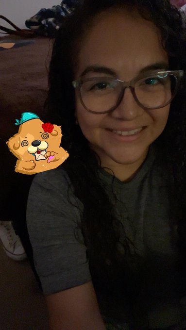 A young woman with glasses and long dark hair smiles warmly for a selfie, wearing a gray t-shirt, with a playful cartoon teddy bear sticker placed over her hand. The image ties to the #RoninMondaySelfie post by showcasing a personal moment meant to inspire kindness and appreciation, while incorporating branded hashtags for @Kind_HQ, @KanstarWorld, @AxieArtGallery, and @RoninSelfie.