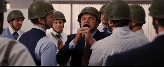 A scene from a film showing Leonardo DiCaprio surrounded by a group of men wearing helmets and business attire, with DiCaprio holding his hands up near his face.