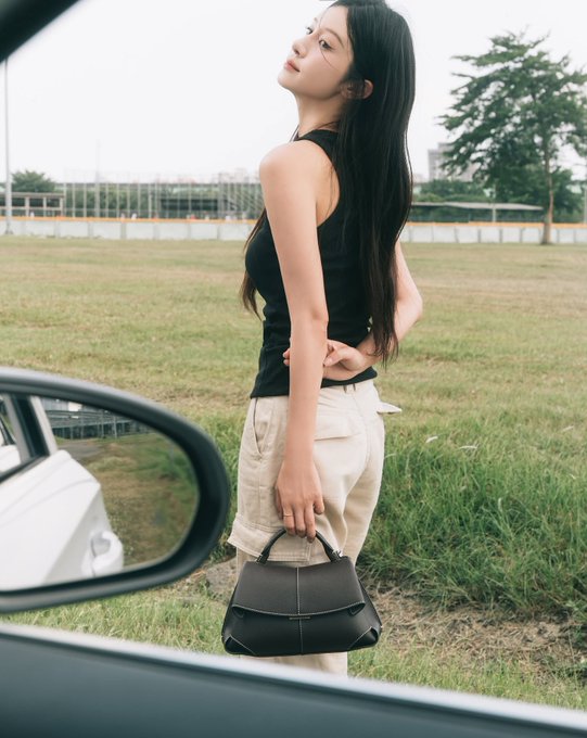 A young woman with long dark hair, wearing a black tank top and beige pants, stands by an open car door holding a black handbag in a grassy field.