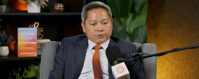 A photograph of an Asian man in a suit and orange tie sitting in a studio with a microphone, with a book titled 'BITCOIN IS FOR EVERYONE' by Natalie Brunell visible in the background.