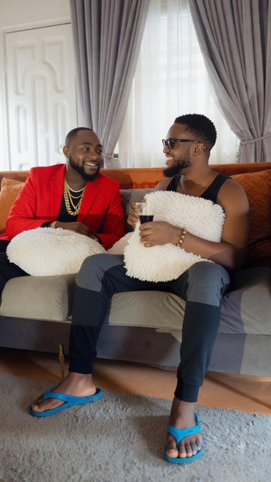 Two men sit on a couch in a living room; one wears a red jacket and laughs while the other, wearing a black tank top and sunglasses, holds a wine glass and smiles.