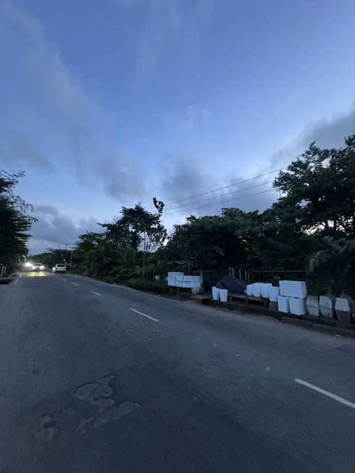 An evening road scene with a cloudy blue sky, trees lining the roadside, and what appears to be white containers or goods stacked along the right side of the road.