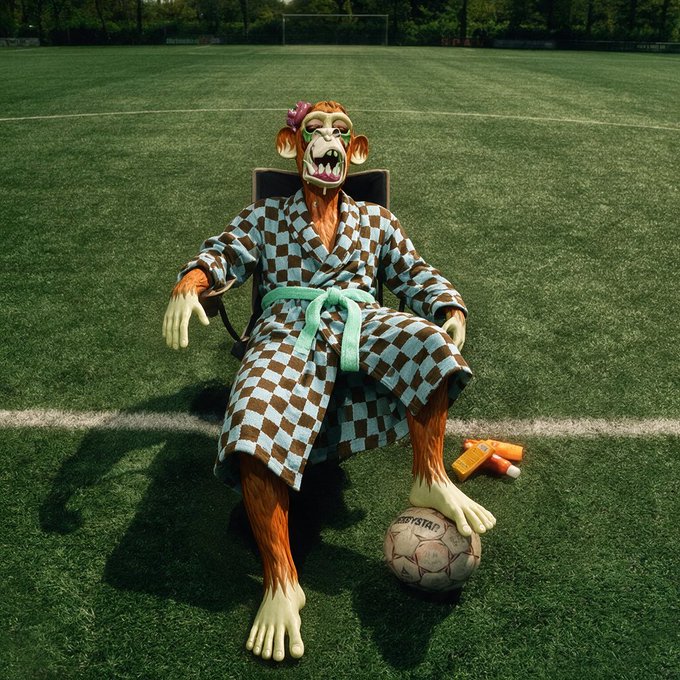 A surreal image of a monkey-like creature wearing a blue and brown checkered robe with a mint green sash, sitting on a chair on a green soccer field with a soccer ball at its feet.