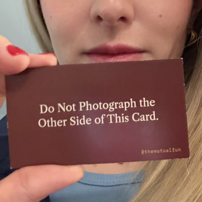 A close-up shows a person with red nail polish holding a brown card that reads 'Do Not Photograph the Other Side of This Card.' with '@themutualfun' in the corner.