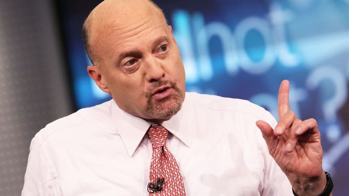 A bald man with a goatee wearing a white shirt and patterned red tie speaks animatedly while pointing his finger upward, with a blurred blue background.