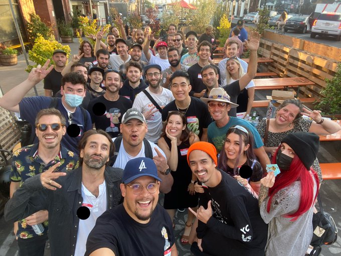 A large group of smiling, cheering people pose for a selfie at an outdoor gathering, with one person wearing an "LA" cap and others flashing peace signs and holding drinks. The "BAYC" reference in the post suggests this is a gathering of Bored Ape Yacht Club NFT community members, likely during one of their real-life meetup events that brought the digital collection's holders together in person.