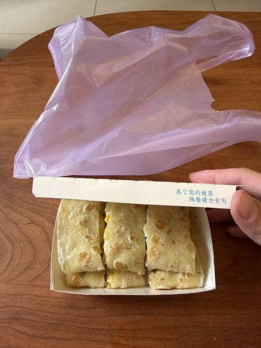 A box of sliced pastries or peanut candies with nuts visible, sitting on a wooden table with a pink plastic bag behind it; the box lid has Chinese text reading '为了您的健康 陰餐請勿食用'.