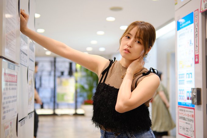 A young Asian woman with brown hair wearing a brown sleeveless top with black fringe detail stands in a Japanese storefront or station, touching her face with one hand while reaching up toward posted notices with the other.
