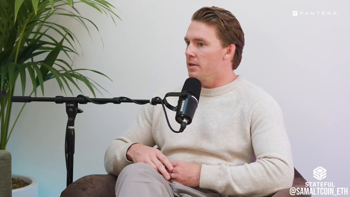 The image shows a man sitting down with his hands folded on his lap while wearing a white sweater. He has a microphone attached to him, suggesting that he might be participating in an interview or a podcast recording session. The background features a plant, adding a touch of nature and life to the scene. The post also mentions the integration of AI into various platforms like World ID, which could potentially impact how people interact with these platforms.