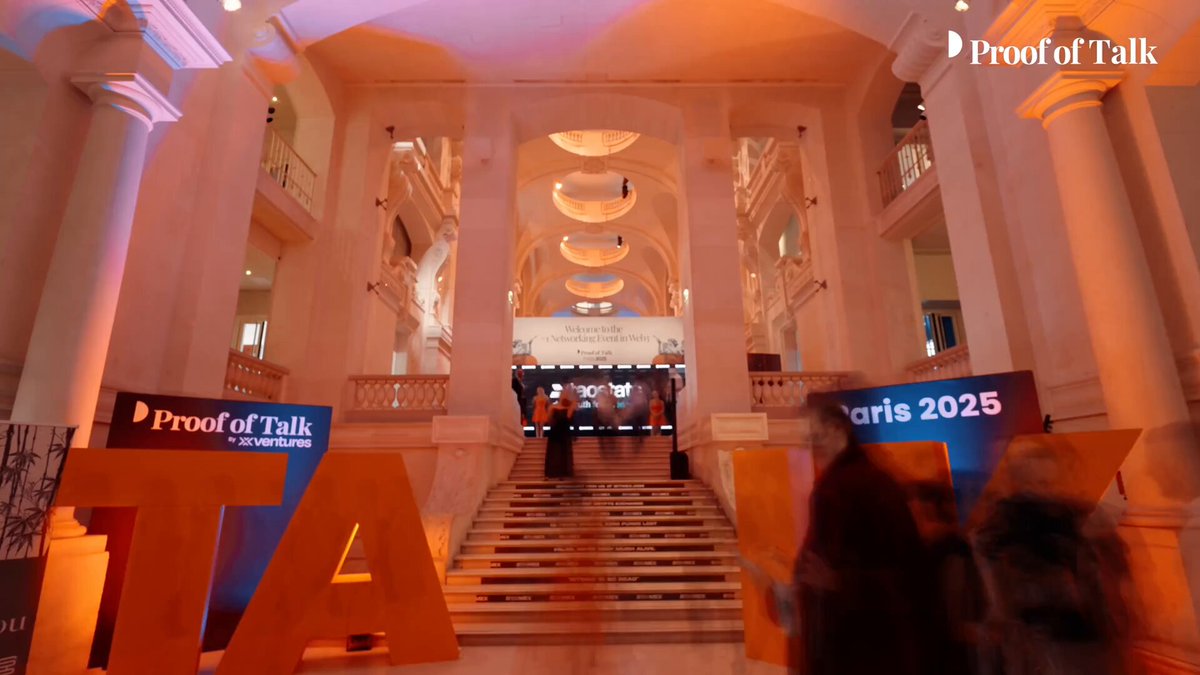The image shows a grand, white building with columns and a large staircase leading up to an entrance. The people inside are gathered around a stage where a presentation or event is taking place. A sign reading "TAO" can be seen near the center of the room. The attendees appear to be engaged in conversation or listening to a speaker on stage. The image also contains text that reads, "Profit of Talk", which could possibly be related to an event or conference happening inside the building.