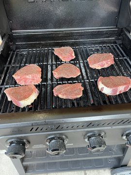 Six raw steaks placed on a black gas grill grates, ready to be cooked, with the grill's control knobs visible in the foreground.