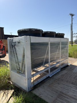 Industrial equipment consisting of black cylindrical tanks stacked on a metal frame wrapped in plastic, sitting on a wooden platform outdoors.