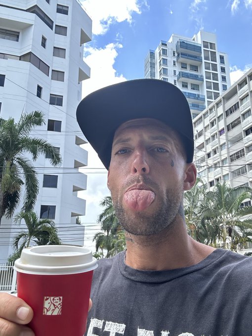 The image features a man wearing a black baseball cap, holding a red coffee cup with white foam on top of his head. The background includes tall buildings, suggesting that he might be in an urban setting or near a city center. The caption "Good morning frens" implies that it is the start of a new day and the man is enjoying his coffee before beginning his daily activities.

The image also contains text elements such as "sa_1296", which could potentially be part of a social media post, an advertisement or some other form of digital content.