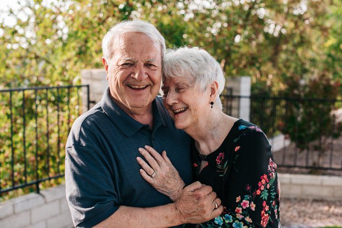 A joyful senior couple embraces warmly outdoors, sharing a laugh together as the post humorously shares their unexpected venture into buying cryptocurrency as potential gifts for their grandchildren.