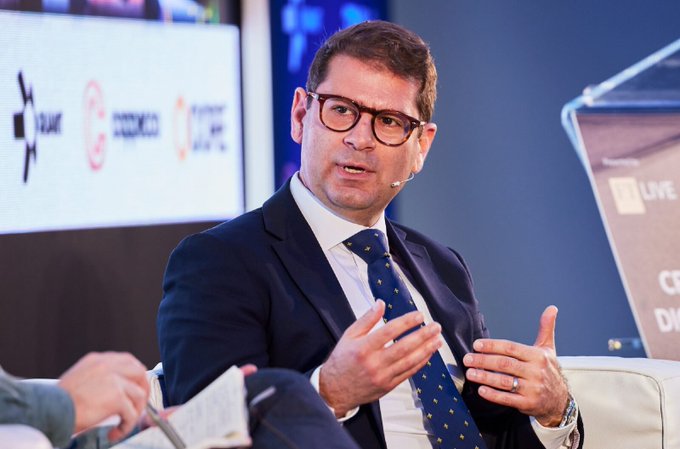 A bespectacled man in a dark suit and dotted tie speaks and gestures while seated on a panel, with a "QUANT" logo visible on the backdrop behind him.