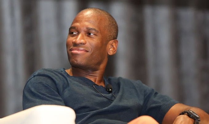A portrait of a smiling bald man in a dark blue t-shirt, appearing to be at an interview or panel discussion.