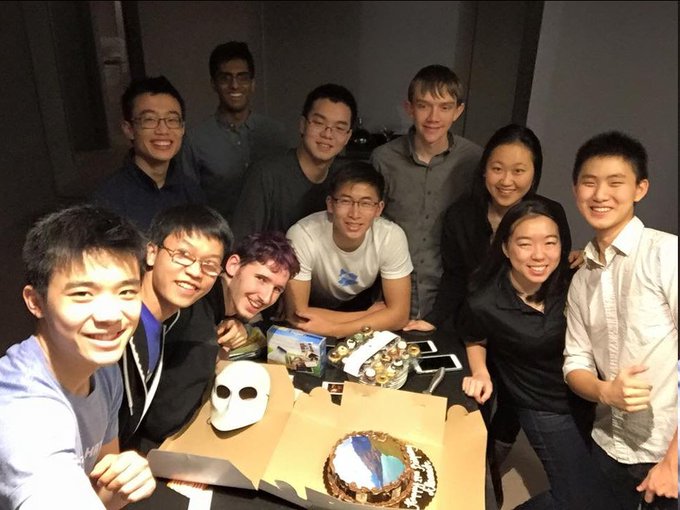 A group of around ten young interns smile together around a table featuring a white mask and a decorated cake, capturing the 2015 HRT intern class referenced in the post that highlights alumni who went on to found major tech companies like Hyperliquid, Scale AI, Cognition, and Decagon.