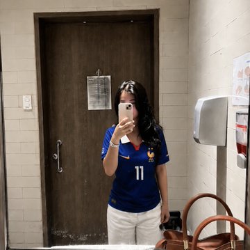 A mirror selfie of a person wearing sunglasses on their head and a blue soccer jersey with the number 11, taking a photo with a smartphone in what appears to be a restroom or hallway.