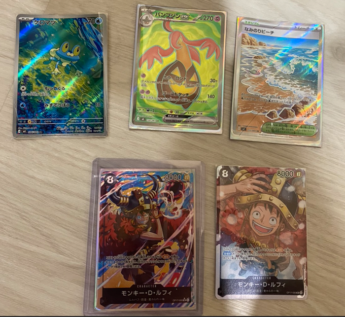 A collection of five Japanese Pokémon trading cards is displayed on a wooden surface, including holographic cards featuring Froakie, Iron Leaves, a beach scene, and two Monkey D. Luffy character cards marked 8000.