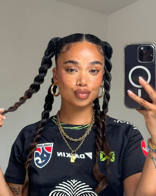 A woman with long braided pigtails, gold necklaces, and hoop earrings takes a mirror selfie with a black phone, dressed in a black shirt featuring a 'THAILAND' and 'PIRELLI' logo patch.