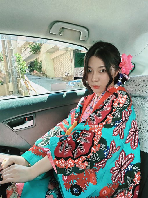 A young woman wearing a vibrant turquoise and red floral kimono with a pink flower hair accessory, sitting in the back seat of a car.