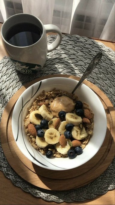A bowl of granola topped with banana slices, blueberries, and almonds sits next to a Starbucks mug of black coffee on a woven placemat.