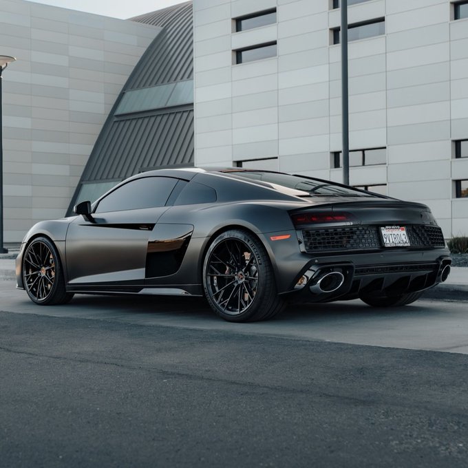 A matte black Audi R8 supercar parked in front of a modern building with curved architecture, featuring bronze/copper accents, custom wheels, and a California license plate.