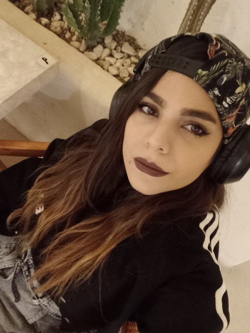 A selfie of a woman with long brown hair, dark lipstick, a backwards floral cap, and headphones, wearing a black Adidas jacket.