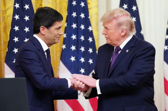 A photograph of two men in suits shaking hands in front of American flags; one man resembles former U.S. President Donald Trump, while the other appears to be a politician or official.