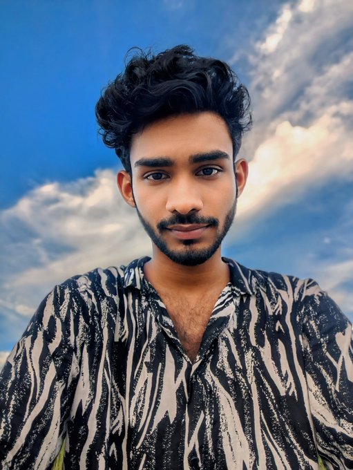 A portrait of a young man with curly black hair and a beard wearing a black and white zebra-striped shirt, photographed against a blue sky with white clouds.