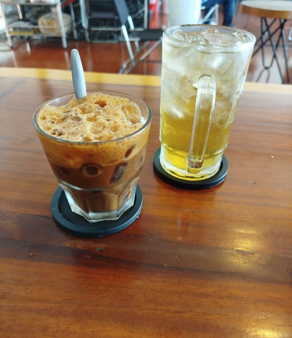 Two iced beverages, an espresso drink with foam and a yellow tea or juice, are served in glass mugs on coasters atop a wooden café table.