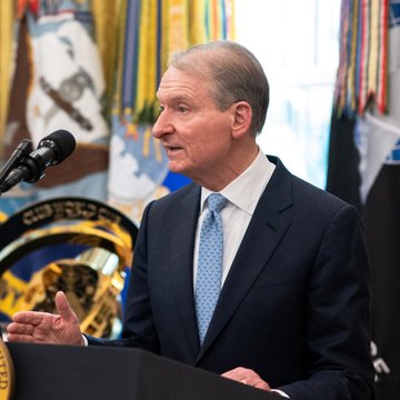 A man in a dark suit and blue tie speaks at a podium with microphones, gesturing with his hand. Flags and a seal are visible in the background.
