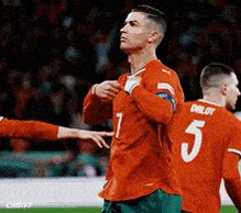 The image shows a soccer player wearing an orange jersey with the number 7 standing on a field, possibly after scoring a goal or celebrating with his teammates. The background of the photo features several people, indicating that this could be a team event or a match celebration. There is also text in the image related to the post about Root and its role as the most boring part of Bittensor for over a year now.