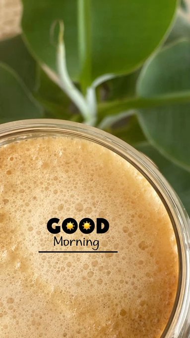 A close-up of a frothy coffee or latte with the text 'GOOD Morning' overlaid, set against a background of green leaves.