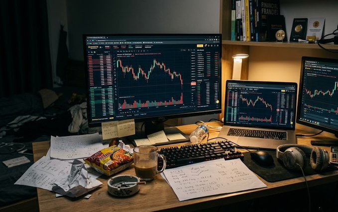 A dimly lit trader's desk setup featuring a large monitor, laptop, and second monitor all displaying trading charts, surrounded by notes, coffee, snacks, and a water bottle.