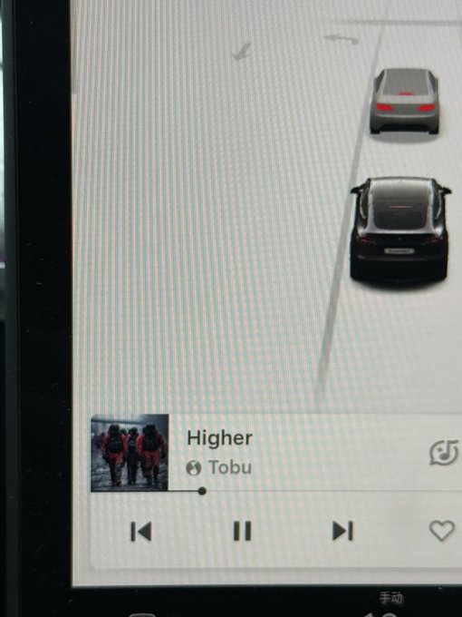 A photo of a car infotainment screen displaying a driving visualization with two vehicles on a road, playing the song 'Higher' by Tobu with standard playback controls visible at the bottom.