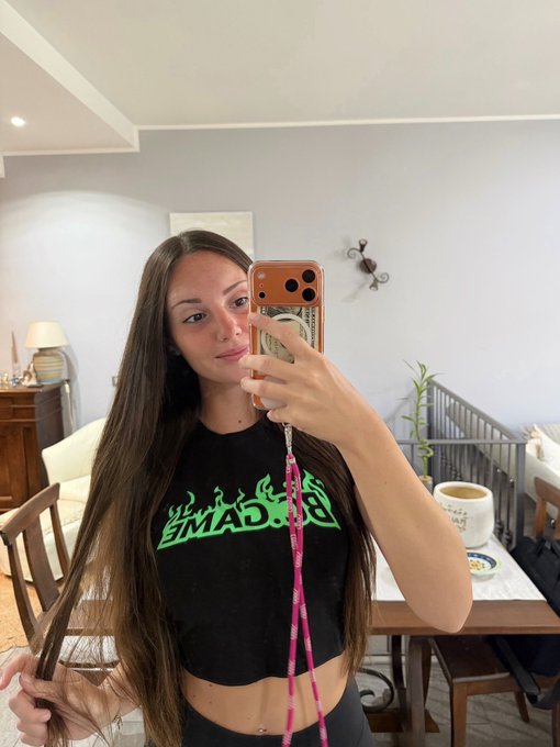 A young woman with long dark hair takes a mirror selfie in a home interior, wearing a black crop top with green graphic text and holding an orange phone with a pink lanyard.