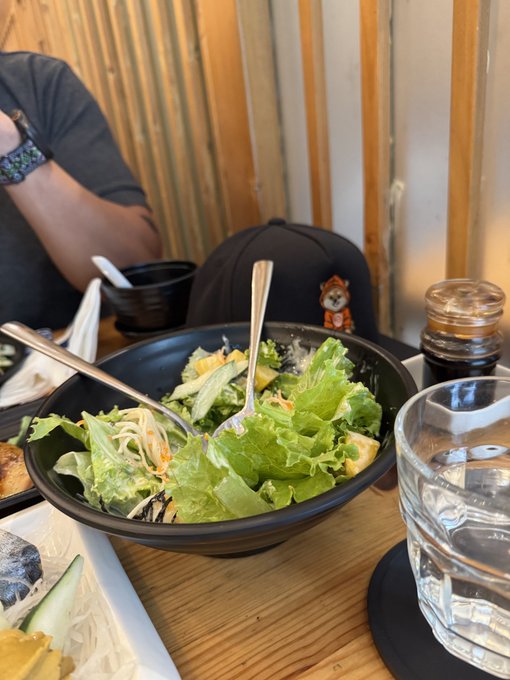 A casual lunch scene featuring a fresh green salad in a black bowl, with a companion across the table wearing a watch. The relaxed dining setting mirrors the post's vibe of "onboarding" a new $DOG supporter over a meal, with a drink of water and sauce bottle suggesting a shared business lunch.