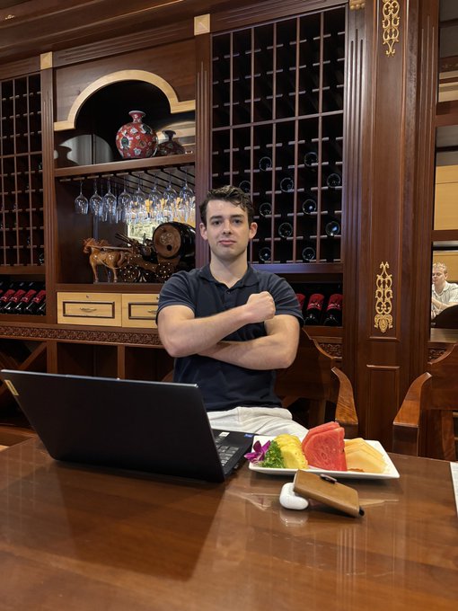 A man in a navy blue polo shirt sits at a wooden table with arms crossed in front of an elaborate dark wood wine cabinet, with a laptop, earbuds case, and a plate of fresh fruit (watermelon, pineapple, orange) on the table.