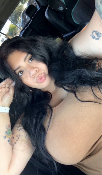 A selfie of a woman with long black hair making a pouty face inside a car, with a tattoo visible on her chest/shoulder area.