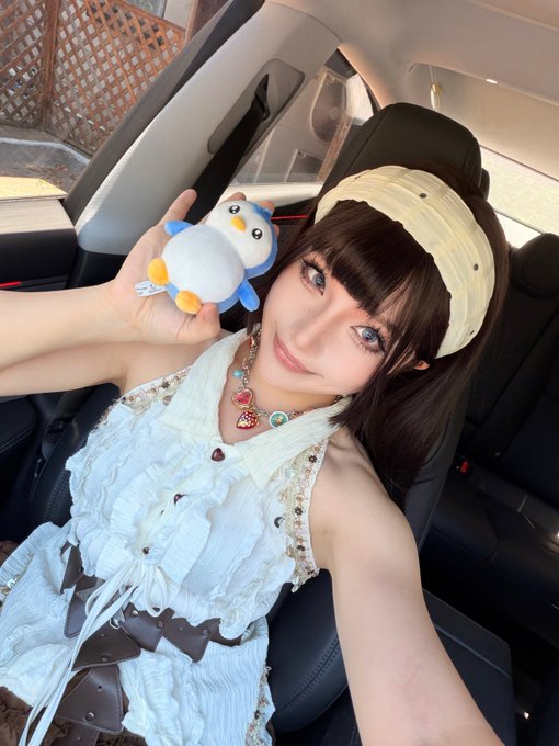A selfie of a young woman with dark hair, wearing a white dress, a beige headband, and colorful charms, holding a small penguin plush toy inside a car.